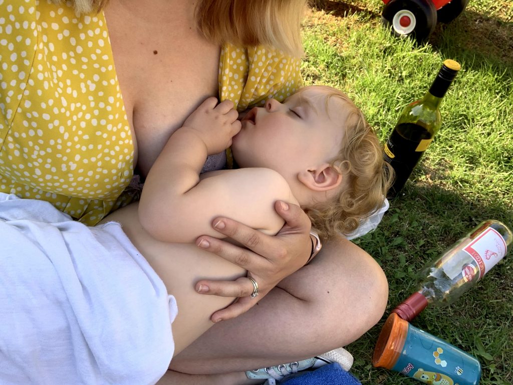  A mum breastfeeding a toddler. The toddler is now asleep, wrapped in white fabric. There are 2 bottles of wine in the shot. 