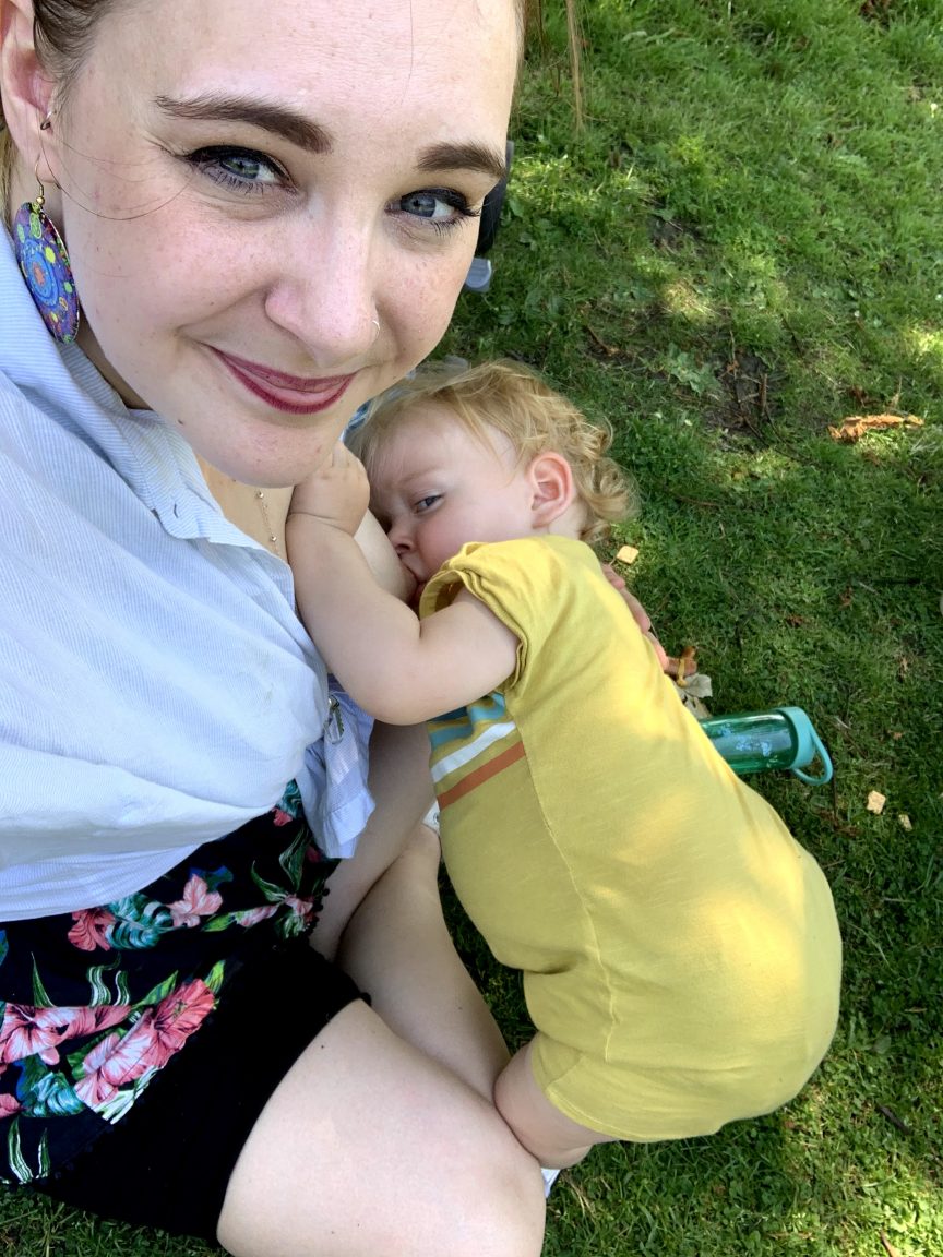 A mum breastfeeding a toddler. The toddler is wearing yellow and is stood up, leaning over.