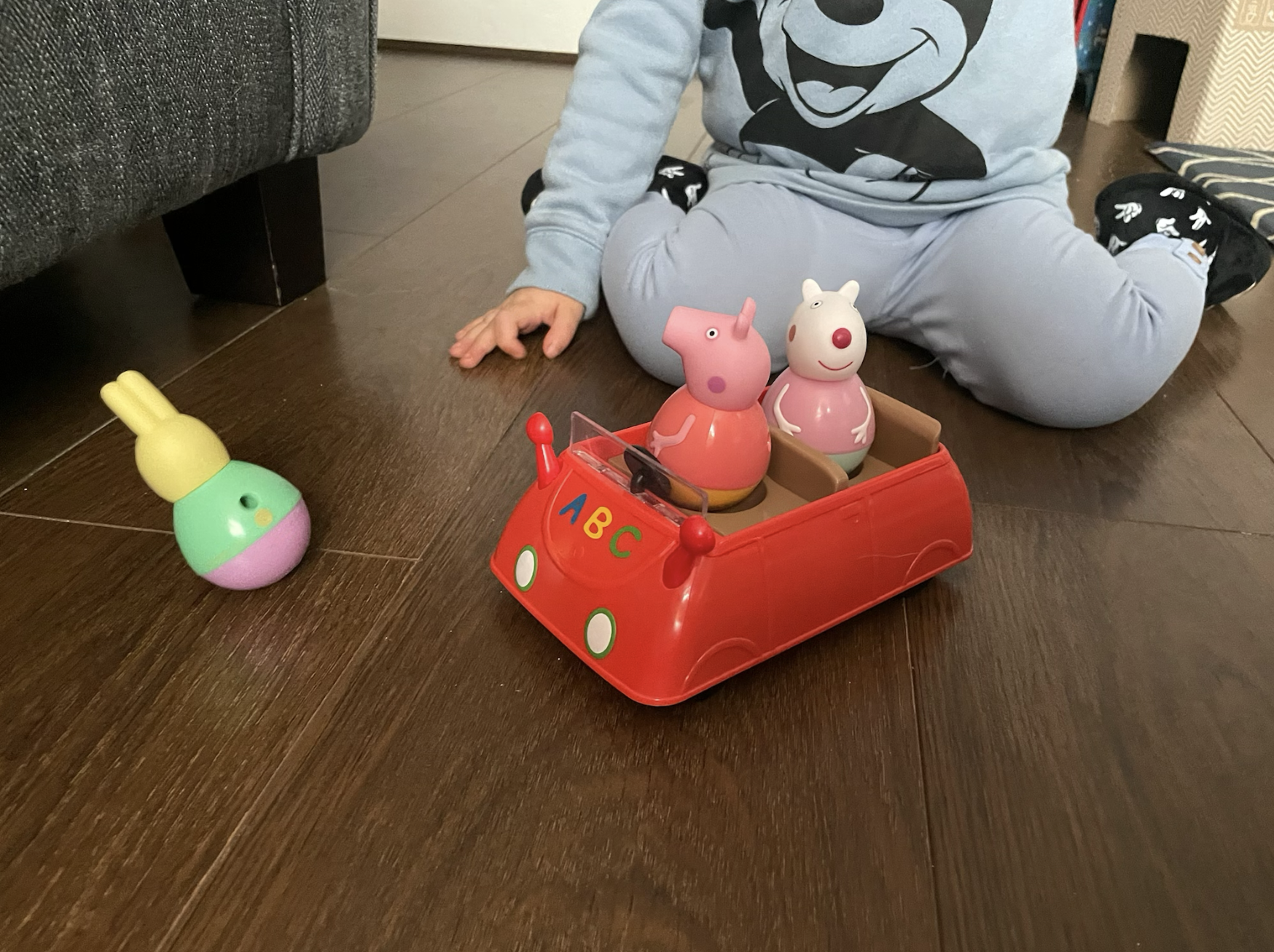 Peppa Pig Weebles, Peppa Pig and Susie Sheep are sat in a red car while Rebecca Rabbit wobbles on a brown wooden floor. A toddlers blue leggings and jumper are in shot playing